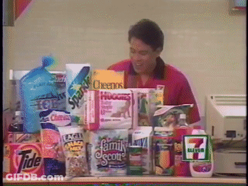 A 7 Eleven Cashier Stands Behind A Counter Full Of Groceries Including Cheerios GIF