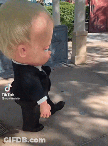 A Baby Doll In A Tuxedo Is Walking On A Sidewalk GIF