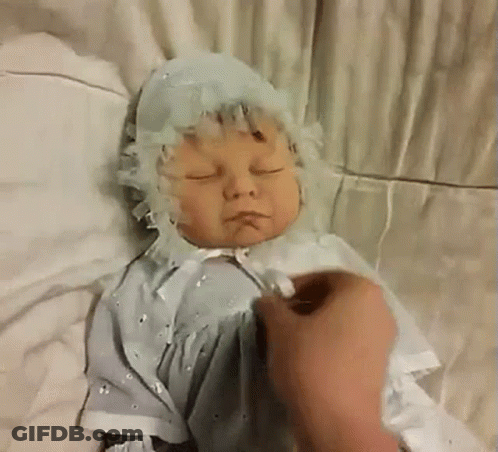A Baby Doll Is Laying On A Bed With Its Eyes Closed And A Person Is Touching It . GIF