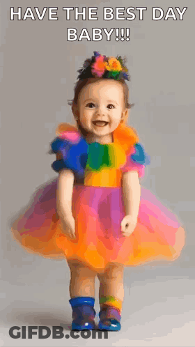 A Baby Girl In A Colorful Dress Is Standing And Smiling . GIF