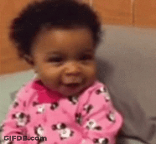 A Baby Girl In A Pink Pajama Set Is Smiling While Sitting On A Bed . GIF