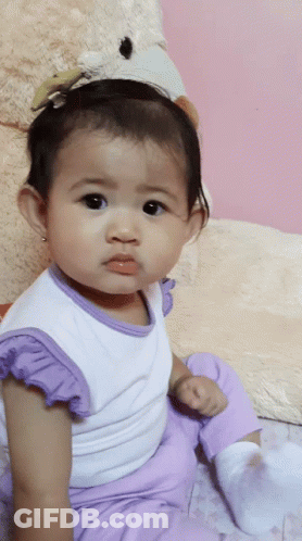 A Baby Girl Wearing Purple Pants And A White Shirt Is Sitting Next To A Teddy Bear GIF