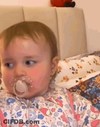 A Baby Girl With A Pacifier In Her Mouth Is Sitting On A Bed . GIF