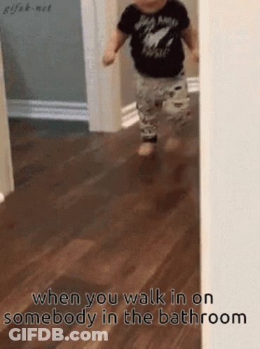A Baby In A Black Shirt Is Walking On A Wooden Floor In A Bathroom . GIF