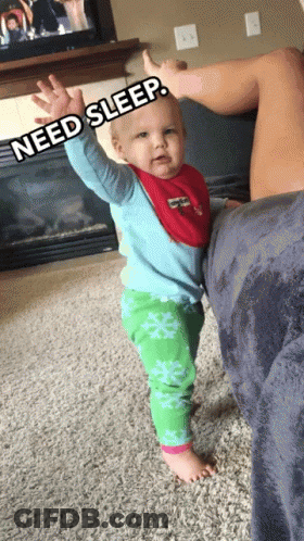 A Baby In A Blue Shirt And Green Pants Is Standing Next To A Woman Who Is Laying On A Couch GIF