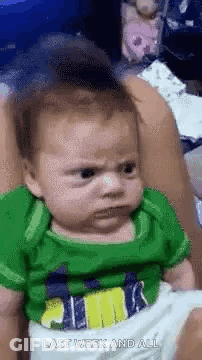 A Baby In A Green Shirt Is Making A Funny Face While Being Held By A Woman . GIF