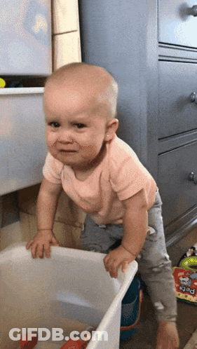 A Baby In A Pink Shirt Is Standing Next To A White Bin With Toys In It GIF