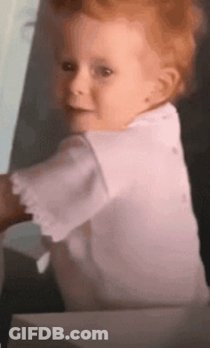 A Baby In A White Shirt Looks At The Camera GIF