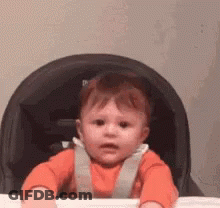 A Baby In An Orange Shirt Is Sitting In A High Chair . GIF