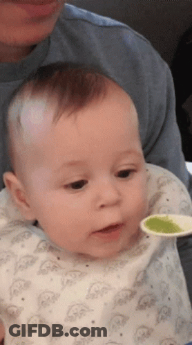 A Baby Is Being Fed With A Spoon By A Man In A Blue Shirt GIF