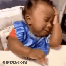 A Baby Is Crying While Sitting At A Table With His Hands On His Head . GIF