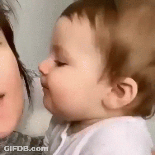 A Baby Is Kissing A Woman On The Cheek And Looking At The Camera . GIF