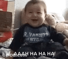 A Baby Is Laughing While Sitting On A Bed With Boxes In The Background . GIF