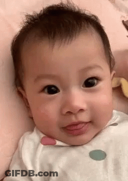 A Baby Is Laying On A Bed And Looking At The Camera With His Tongue Out . GIF