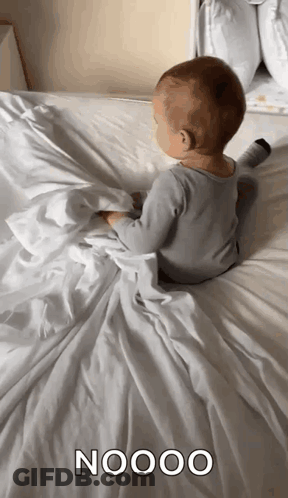 A Baby Is Laying On A Bed With A White Blanket And The Word Nooo Written On It GIF