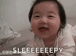 A Baby Is Laying On Its Stomach On A Bed And Smiling With Its Tongue Out . GIF