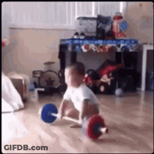 A Baby Is Lifting A Barbell On The Floor In A Room . GIF