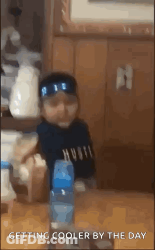 A Baby Is Sitting At A Table With A Bottle Of Water In Front Of Him . GIF
