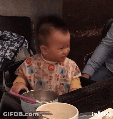 A Baby Is Sitting At A Table With A Bowl Of Food And A Spoon And Fork GIF