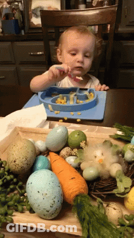 A Baby Is Sitting At A Table With A Plate Of Food And A Spoon GIF