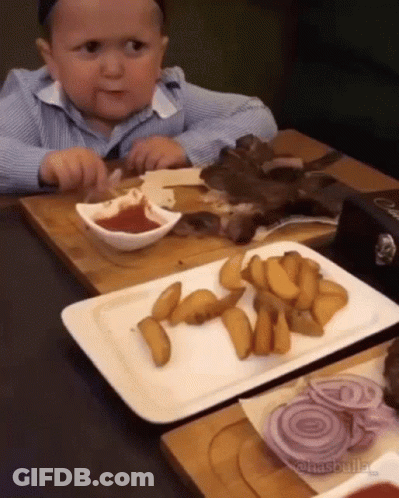 A Baby Is Sitting At A Table With A Plate Of French Fries And A Plate Of Meat . GIF