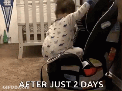 A Baby Is Sitting In A Car Seat Next To A Crib In A Bedroom . GIF