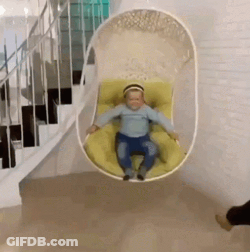 A Baby Is Sitting In A Hanging Chair In A Room Next To Stairs . GIF
