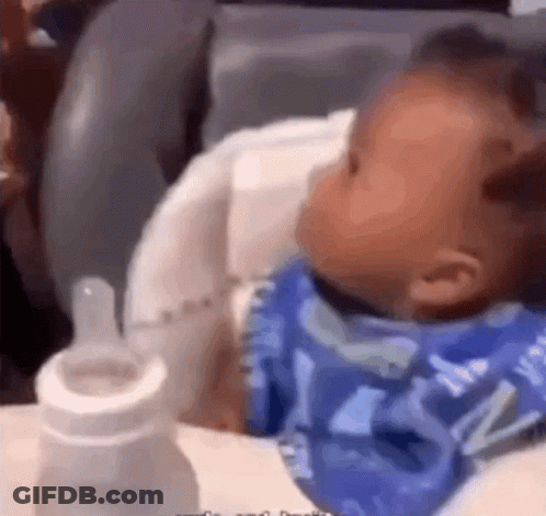 A Baby Is Sitting In A High Chair With A Bottle GIF