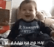 A Baby Is Sitting On A Bed And Laughing While Wearing A Varsity Shirt . GIF