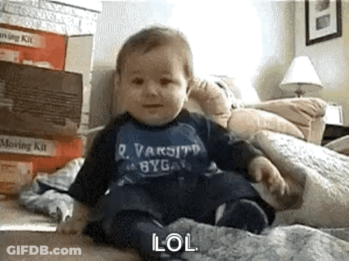A Baby Is Sitting On A Bed Wearing A Blue Shirt That Says Varsity GIF