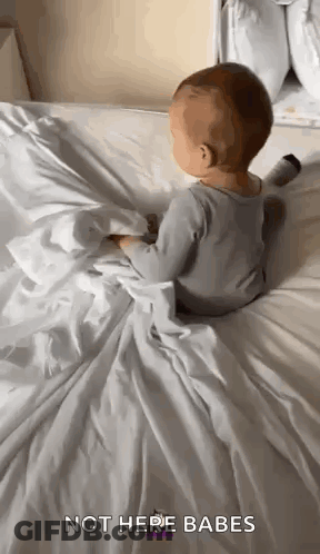 A Baby Is Sitting On A Bed With A White Blanket And A Blanket . GIF