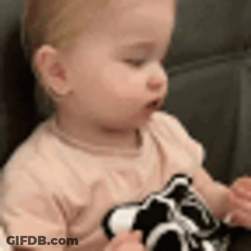A Baby Is Sitting On A Couch Holding A Black And White Toy . GIF
