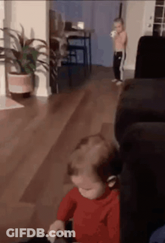 A Baby Is Sitting On A Couch In A Living Room While A Little Boy Stands In The Background . GIF