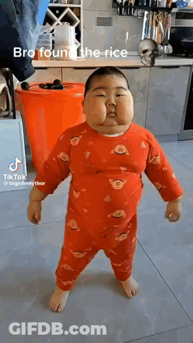 A Baby Is Standing In A Kitchen Wearing A Red Pajama Set . GIF