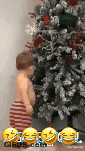 A Baby Is Standing In Front Of A Christmas Tree With Tears Running Down His Face . GIF