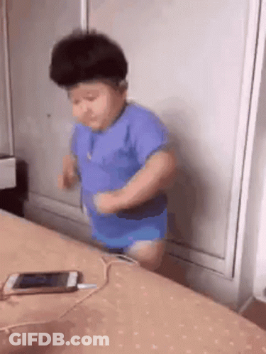 A Baby Is Standing On A Bed Next To A Cell Phone . GIF