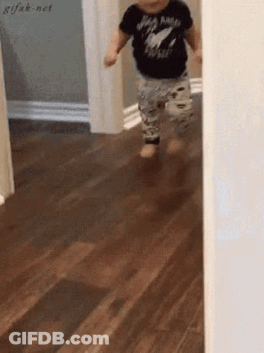 A Baby Is Walking On A Wooden Floor Wearing A Black Shirt That Says Rock N Roll GIF