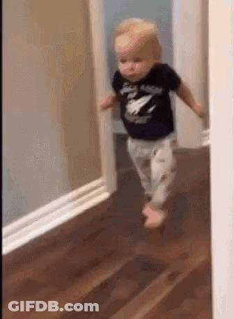 A Baby Is Walking Through A Doorway On A Hardwood Floor . GIF
