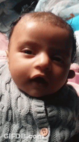 A Baby Is Wearing A Grey Sweater With A Button On It GIF