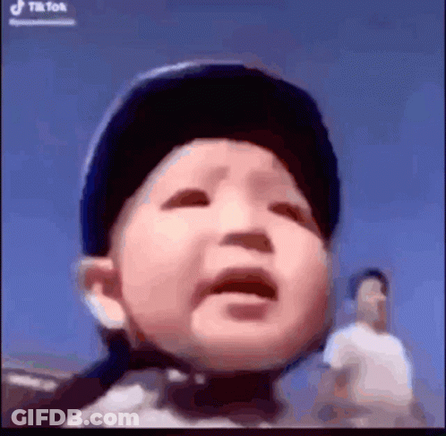 A Baby Is Wearing A Helmet And Crying While A Man Stands Behind Him . GIF
