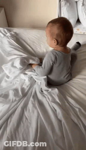A Baby Laying On A Bed With A White Blanket GIF