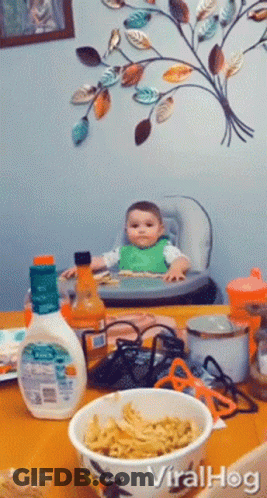A Baby Sits In A High Chair Next To A Bowl Of Noodles GIF