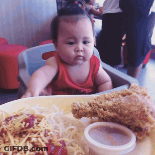 A Baby Sitting In A High Chair With A Plate Of Food In Front Of Her GIF