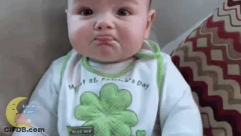 A Baby Wearing A Bib With A Clover On It Is Making A Funny Face While Sitting On A Couch . GIF