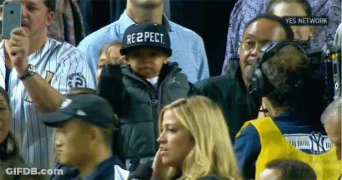 A Baby Wearing A Hat That Says Respect Stands In A Crowd GIF
