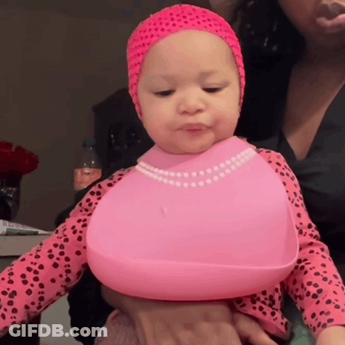 A Baby Wearing A Pink Bib And A Pink Headband Is Being Held By A Woman GIF