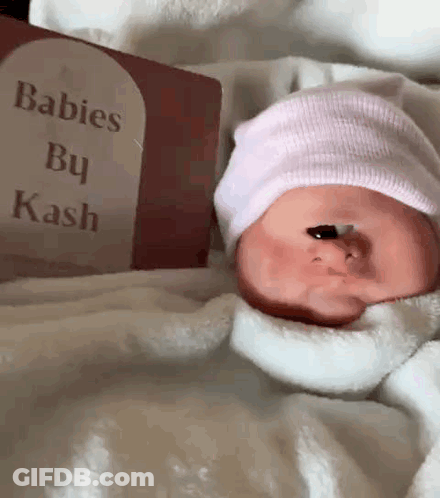 A Baby Wearing A Pink Hat Is Laying Next To A Book Titled Babies By Kash GIF