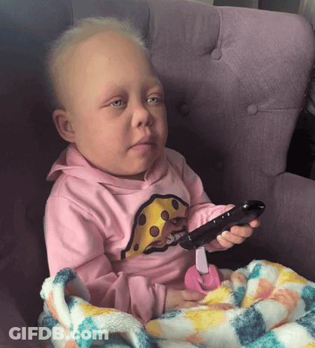 A Baby Wearing A Pink Shirt With Cheese On It Holds A Remote Control GIF