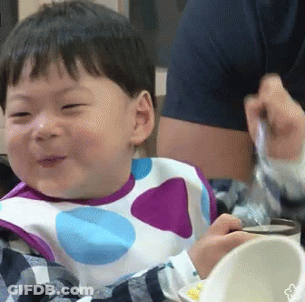A Baby Wearing A Polka Dot Bib Is Smiling While Eating From A Bowl . GIF