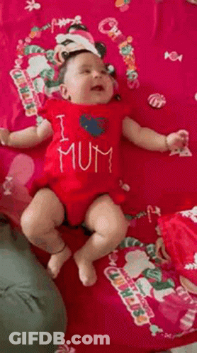 A Baby Wearing A Red Shirt That Says `` I Love Mum '' Is Laying On A Pink Blanket . GIF